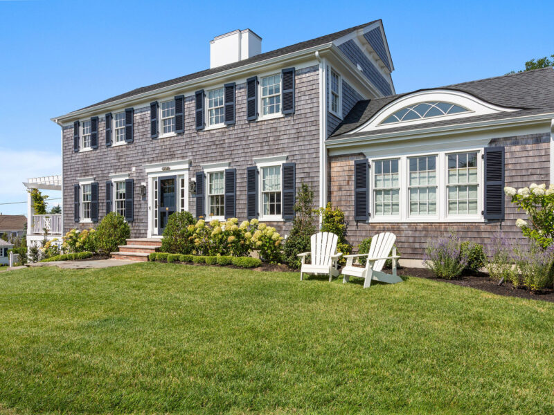 A two-story gray shingle house with white trim and black shutters on a manicured lawn – McPhee Associates of Cape Cod.